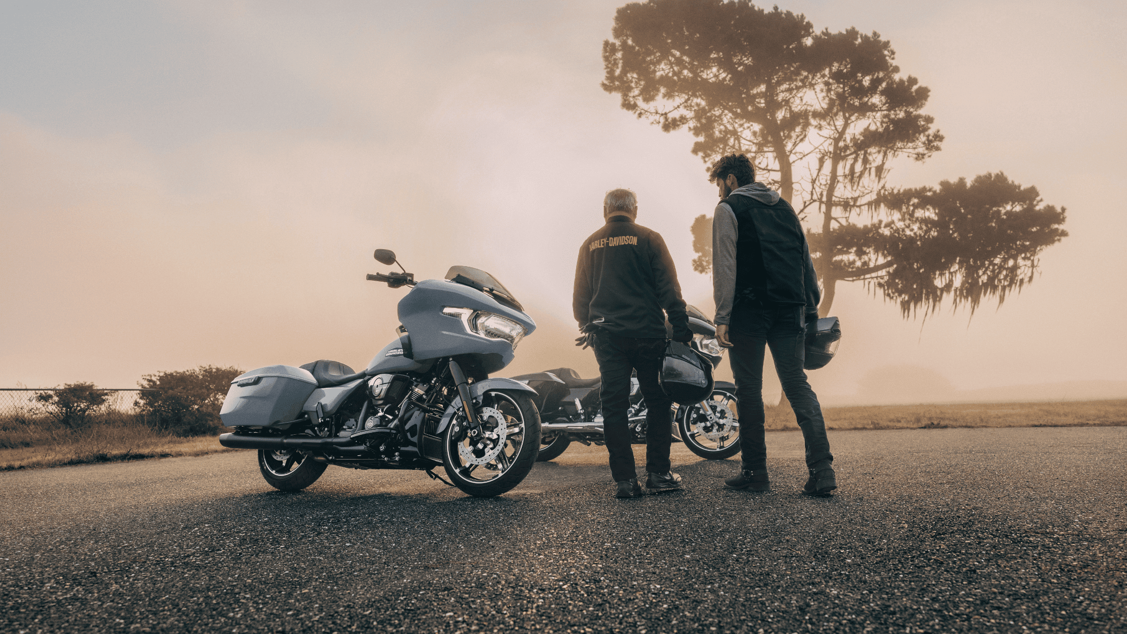 A pair of people standing next to a 2024 Harley-Davidson® Road Glide® motorcycle along a picturesque road.