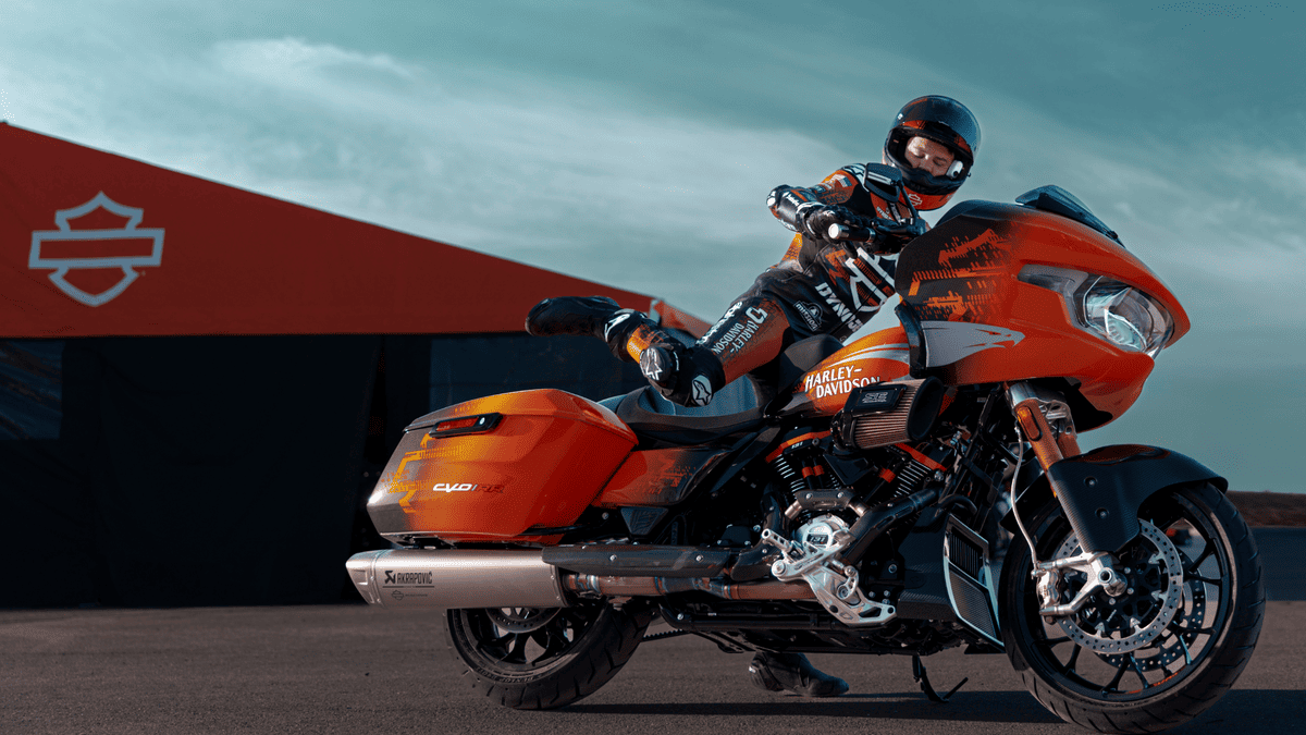 Rider mounts the bold orange 2025 Harley-Davidson® CVO™ Road Glide® RR near Bellaire, TX, in front of a Harley-Davidson® tent with racing gear on.